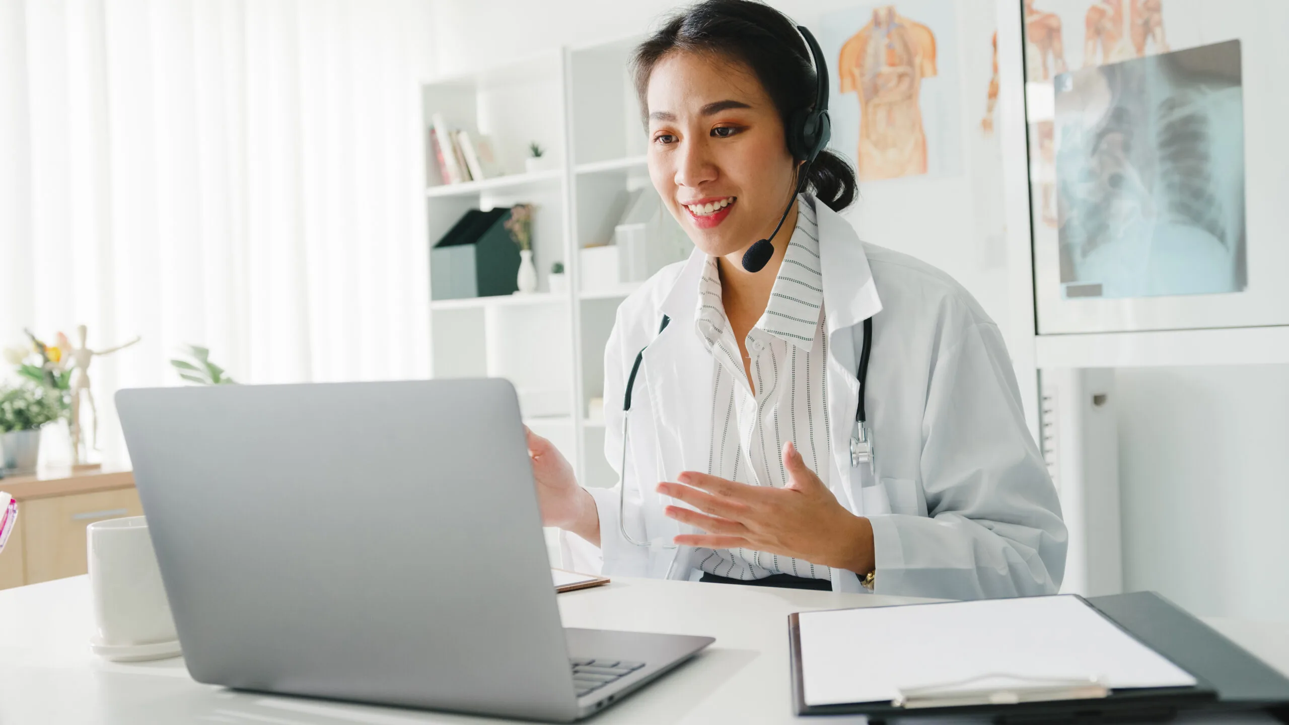 Young Asia lady doctor in white medical uniform with stethoscope using computer laptop talking video conference call with patient at desk in health clinic or hospital. Consulting and therapy concept.